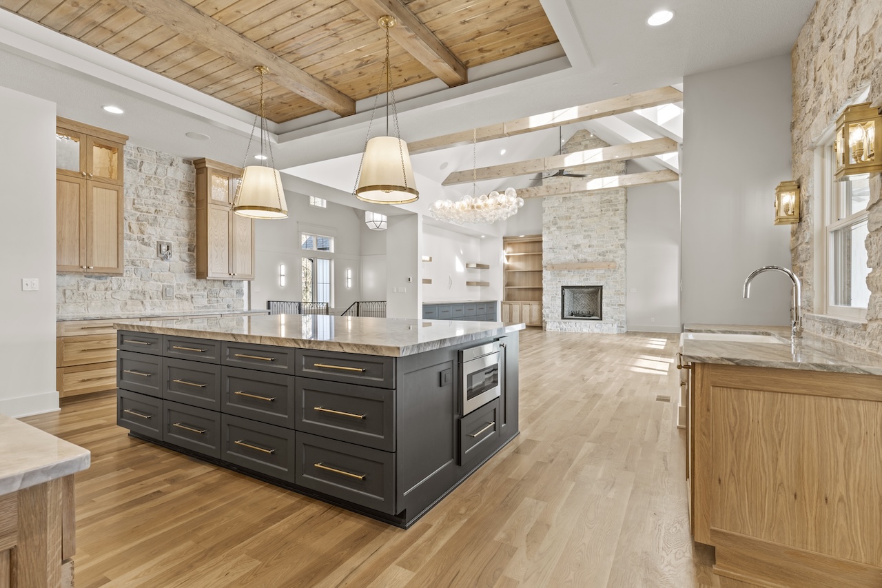 Open-concept kitchen featuring a large dark gray island with marble countertop, brass hardware, and wooden cabinetry against stone walls with pendant lights.